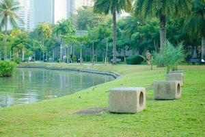serene park scene featuring stone benches by tranquil lake, surrounded by lush greenery and palm trees. People enjoy leisure activities in background, creating peaceful atmosphere photo