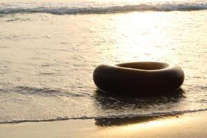 black inflatable tire floats on calm beach water during sunset, creating serene atmosphere. gentle waves reflect warm golden light, enhancing tranquil scene photo
