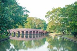 serene park scene featuring pink bridge over calm waterway, surrounded by lush green trees and vibrant foliage, creating peaceful atmosphere photo