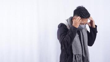 Young Asian Man Wearing Jacket, Scarf, and Mask Suffering from Bad Headache on White Background photo
