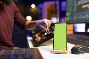 Sound engineer uses controls and amplifier on mixing console to create beautiful sound effects and adjust volume levels on tracks. Producer using greenscreen display on phone, editing soundtracks. photo