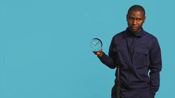 Security guard holding clock, rushing people out of property, isolated over studio backdrop. Man in uniform showing visitors time is passing, preparing for closing down location, camera B photo