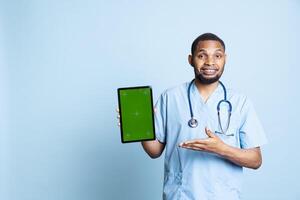 Portrait of male nurse showing medical instructions on tablet with green screen, pointing at device. Hospital employee presenting chroma key display, standing against blue background. photo