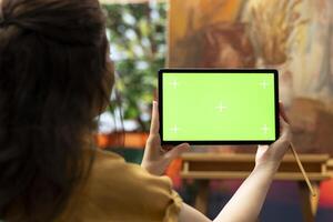 Painter holding a device with isolated mockup screen in her art studio, looking at her finished masterpiece with oil paints on canvas. Beautiful rustic artwork for a fine art gallery, creativity. photo