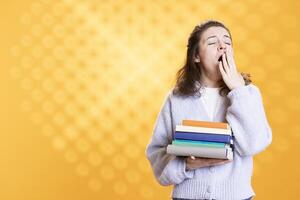 Tired woman yawning while holding books conveying boredom of reading concept, studio background. Exhausted person with novels in arms showing dissatisfaction for tedious literature photo