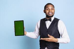 Doorkeeper bellboy poses with green screen on tablet at the hotel reception, working in hospitality industry and greeting clients. Lodging resort employee poses against blue background. photo