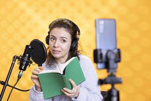 Influencer looking at phone camera, filming herself reading book, creating audiobook, isolated over studio background. Content creator doing vlog while producing digital recording on novel photo