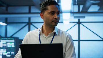 System administrator in data center using notebook to design, implement and manage gear, ensuring optimal performance and reliability. Indian man in server room coding on laptop, camera A photo