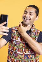 Cheerful man using cellphone to take selfies, rubbing chin. Smiling social media user taking photos using phone selfie camera, doing ego stroking hand gesturing, studio background