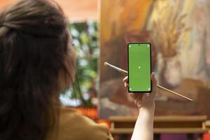 Painter holding a device with isolated mockup screen in her art studio, looking at her finished masterpiece with oil paints on canvas. Beautiful rustic artwork for a fine art gallery, creativity. photo