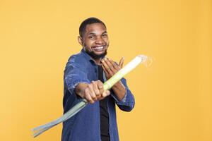 African american funny fighter pretending to do a combat move with a leek, holding green onion as a bat against yellow background. Guy fooling around and having fun with zero waste produce. photo
