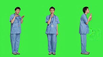 Medical assistant asking to keep quiet and be silent against greenscreen, showing hush mute symbol to keep privacy and silence. Young nurse showing secrecy taboo sign. Camera A. photo