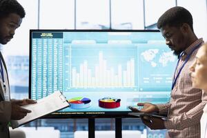 Team of workers presenting infographics and sales numbers on an interactive board, boardroom activity. Diverse group of professionals engages in a project management meeting for growth. photo