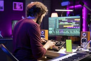 Music engineer twisting pre amp knobs on mixing console in home studio, having phone with greenscreen layout. Composer musician learning to edit soundtracks with daw software. photo