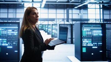 Woman walking in high tech facility, using laptop to patch network issues leading to slow data transfer rates. Employee in data center fixing errors affecting servers performance, camera B photo