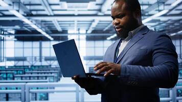Computer scientist in server hub checking recovery plan on laptop, monitoring hardware energy consumption. IT specialist uses notebook in data center facility, ensuring rigs are functioning, camera B photo