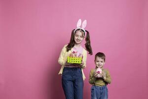 Sweet children holding festive easter decorations on camera, posing with confidence and feeling happy about spring festivity. Brother and sister showing their handmade craft of april ornaments. photo