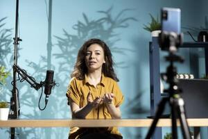 Woman recording livestream in studio, using cellphone selfie camera on tripod and mic to ensure crisp sound and quality. Online star speaking into professional microphone, discussing with fans photo