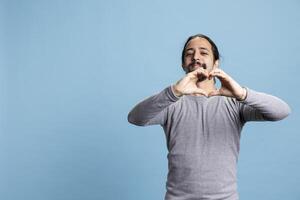 Romantic person showing a heart shape sign against blue background, doing a symbol of affection and passion in studio. Young adult acting playful in the art of seduction on camera. photo