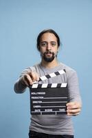 Middle eastern guy holding a filming slate movie production to cut a scene, working in cinematography as a filmmaker against blue background. Cameraman showing the clapper board in studio. photo