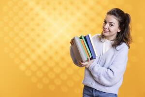 Portrait of cheerful woman carrying stack of textbooks, gathering information for school exam, isolated over studio background. Smiling pupil holding pile of books, preparing for university assessment photo