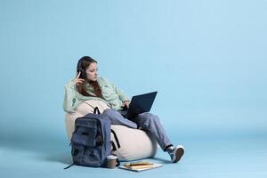 Woman scrolling on laptop to check social media feed and listening music, relaxing on beanbag. Girl wearing headphones, browsing internet websites on notebook, isolated over blue studio background photo