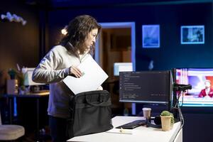 Woman packing laptop after finishing doing software quality assurance and reading source code. Engineer placing notebook in backpack after inspecting coding on desktop PC and finding all issues photo