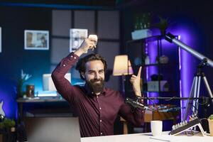 Smiling influencer using light bulb to illustrate idea, discovering brilliant innovation to tell audience. Happy media star pointing finger up to show inspiration concept while filming in apartment photo