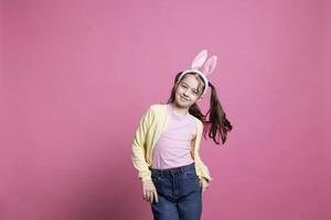 Adorable young kid dancing around in front of camera, posing with confidence and having fun with music. Little child being cheerful with dance moves over pink background, bunny ears and pigtails. photo