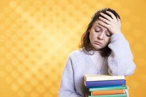 Woman with worried facial expression doing face palm from migraine pain, carrying books for school exam, studio background. Stressed student with textbooks suffering headache from too much homework photo