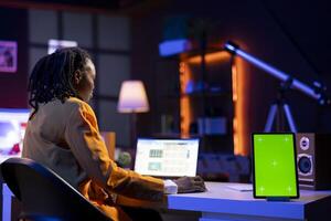 Green screen mockup tablet next to remote employee completing spreadsheets on laptop. Chroma key isolated screen device next to worker in home office editing documents on notebook photo