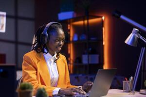 Remote worker listening music, analyzing data on laptop at desk, compiling analytical reports. Woman in home office wearing headphones, using notebook device, typing information photo