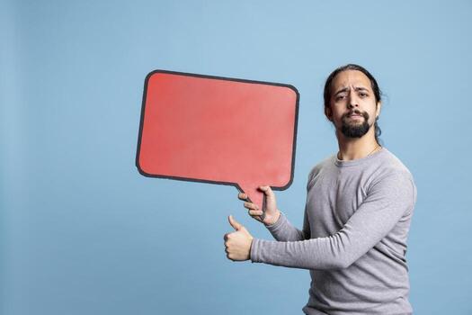 Confident person holds the red cardboard speech bubble on camera, doing an advertisement with a friendly positive attitude. Young smiling man presenting the empty billboard icon in studio. photo