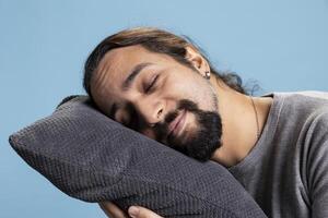 Sleepy young man taking a nap with a pillow standing against blue background, pretending to fall asleep on camera. Exhausted person dealing with burnout and fatigue, tired male model. photo