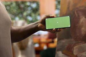 Young artist looking at isolated chroma key display in his painting atelier, using device next to canvas with a colorful masterpiece. Painter creating a rustic artwork for the gallery exhibition. photo