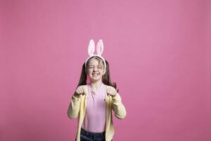 Sweet schoolgirl with bunny ears jumping in studio, impersonating a rabbit for easter holiday festivity. Young cheerful child bouncing around against pink background, acting silly and funny. photo