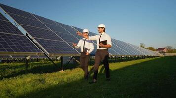 Two investors in helmets and formal attire walk along a solar panel station, discussing the project's performance and future development video