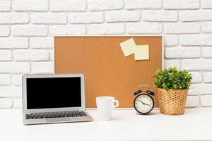Workspace setup featuring a laptop, plant, clock, and corkboard in a bright, minimalistic environment photo