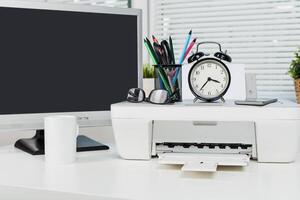 Office workspace featuring printer, clock, stationery, and computer set up during daytime hours photo