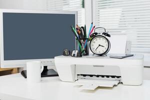 Desk setup with printer, clock, and computer in a bright office environment during daytime work hours photo