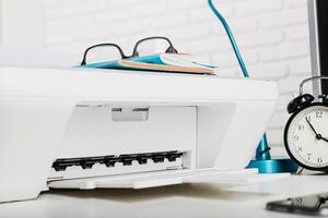 Office workspace with printer, glasses, books, and clock resting on a desk during the day photo