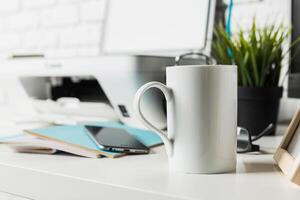 Coffee mug placed on a desk next to a smartphone, printer, and notebook during a work session in a cozy office space photo