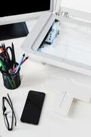Office workspace with a printer, smartphone, stationery, and glasses on a desk during the day photo