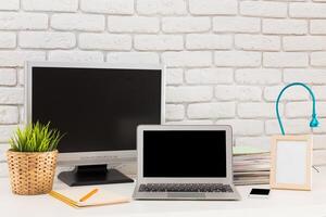 Workstation setup with a laptop, monitor, and stationery on a desk against a white brick wall in a well-lit space photo