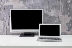 Computer setup with desktop monitor and laptop on a minimalist white table against a textured gray wall photo