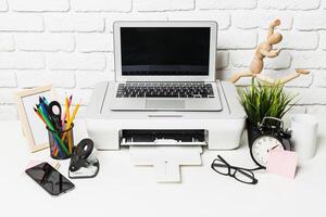 Office workspace with laptop, printer, and stationary on a white brick wall background during working hours photo