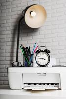 Workspace with a printer, clock, and stationary under a lamp on a textured wall during the day photo