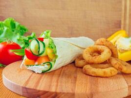 Fresh vegetable wrap served with crispy onion rings on a wooden cutting board in a cozy kitchen setting photo