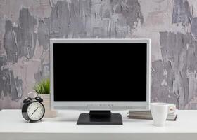 Modern workspace setup featuring a blank monitor, clock, plant, and coffee cup on a desk with textured wall background photo