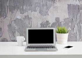 Modern workspace featuring a laptop, smartphone, coffee mug, and potted plant on a white desk with textured gray wall backdrop photo
